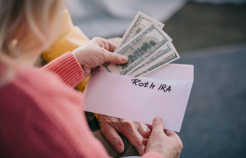 A close-up of an elderly couple holding an envelope labeled "Roth IRA" and counting U.S. dollar bills, symbolizing retirement savings or early withdrawal from a retirement account.