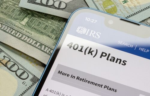 Smartphone displaying IRS website page on 401(k) retirement plans, placed on top of scattered U.S. $100 bills.