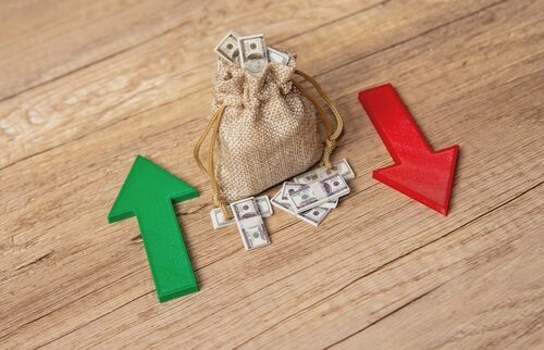 Burlap sack filled with dollar bills placed between a large green upward arrow and a large red downward arrow on a wooden surface, symbolizing financial growth and decline.