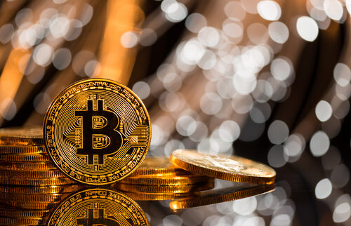 A stack of shiny gold Bitcoin coins with the Bitcoin symbol prominently displayed, set against a blurred background of glowing white and golden lights.