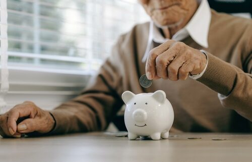Senior person placing a coin into a white piggy bank, symbolizing retirement savings and planning for longevity with a self-directed IRA.